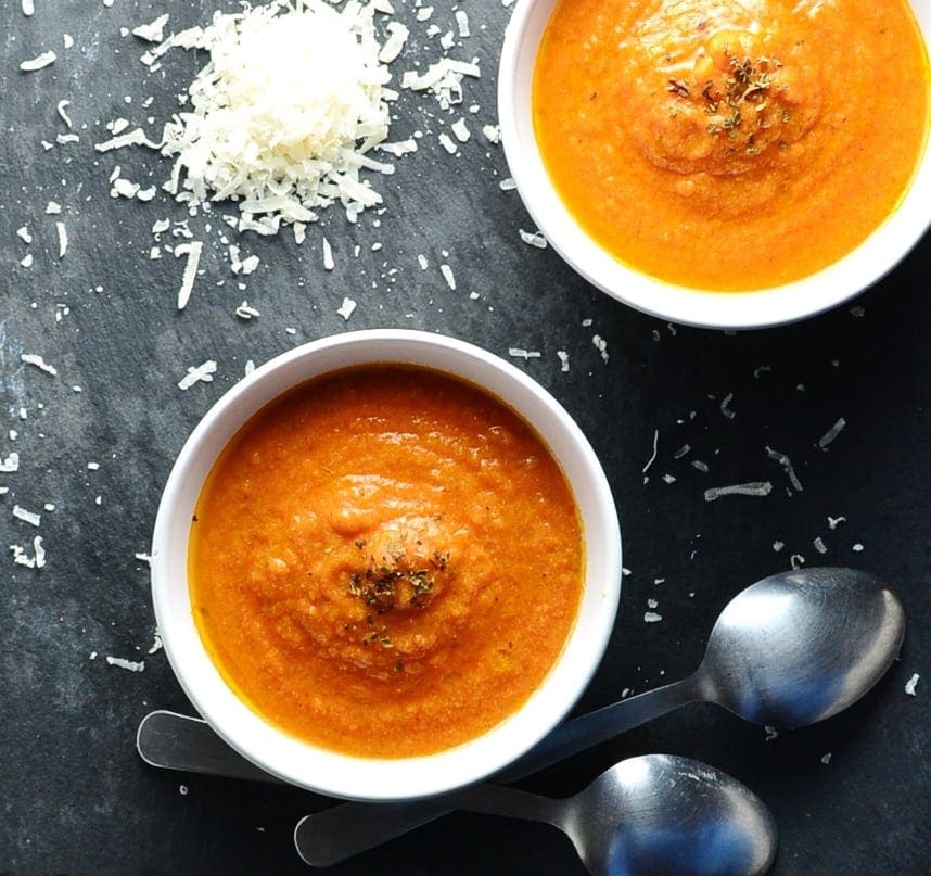 Top down view of creamy tomato soup in 2 white bowls with grated cheese and 2 spoons on top of dark surface.