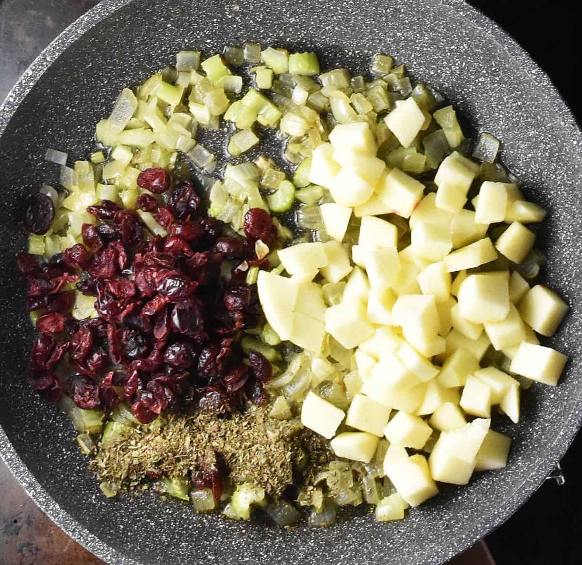 Chopped apple, dried cranberries, herbs and sauteed onion in large grey pan.