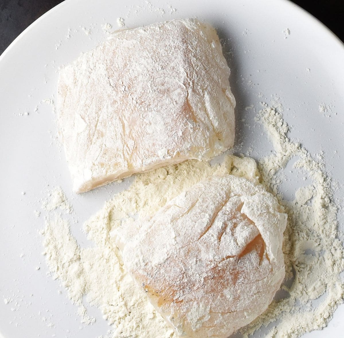 Top down view of 2 pieces of fish coated in flour on white plate.