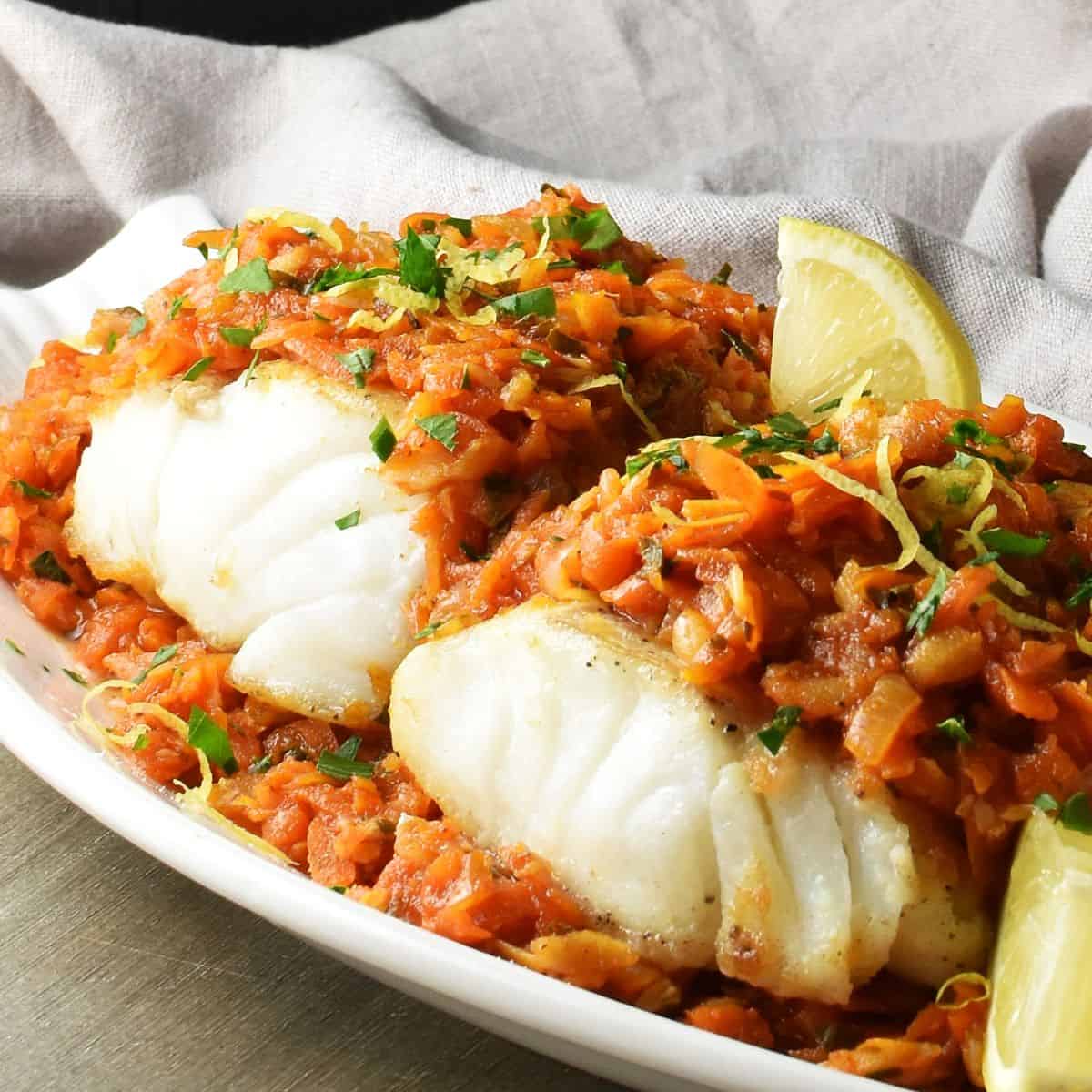 Side view of Polish fish coated in thick vegetable sauce on white plate with lemon wedges.