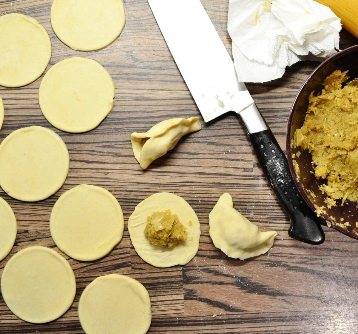 Forming sauerkraut pierogi with round dough shapes, sauerkraut mixture in bowl and knife on wooden surface.