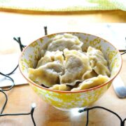 Sauerkraut pierogi in yellow-and-white patterned bowl with Christmas themed coasters, Christmas lights and green cloth in background on light wooden surface.
