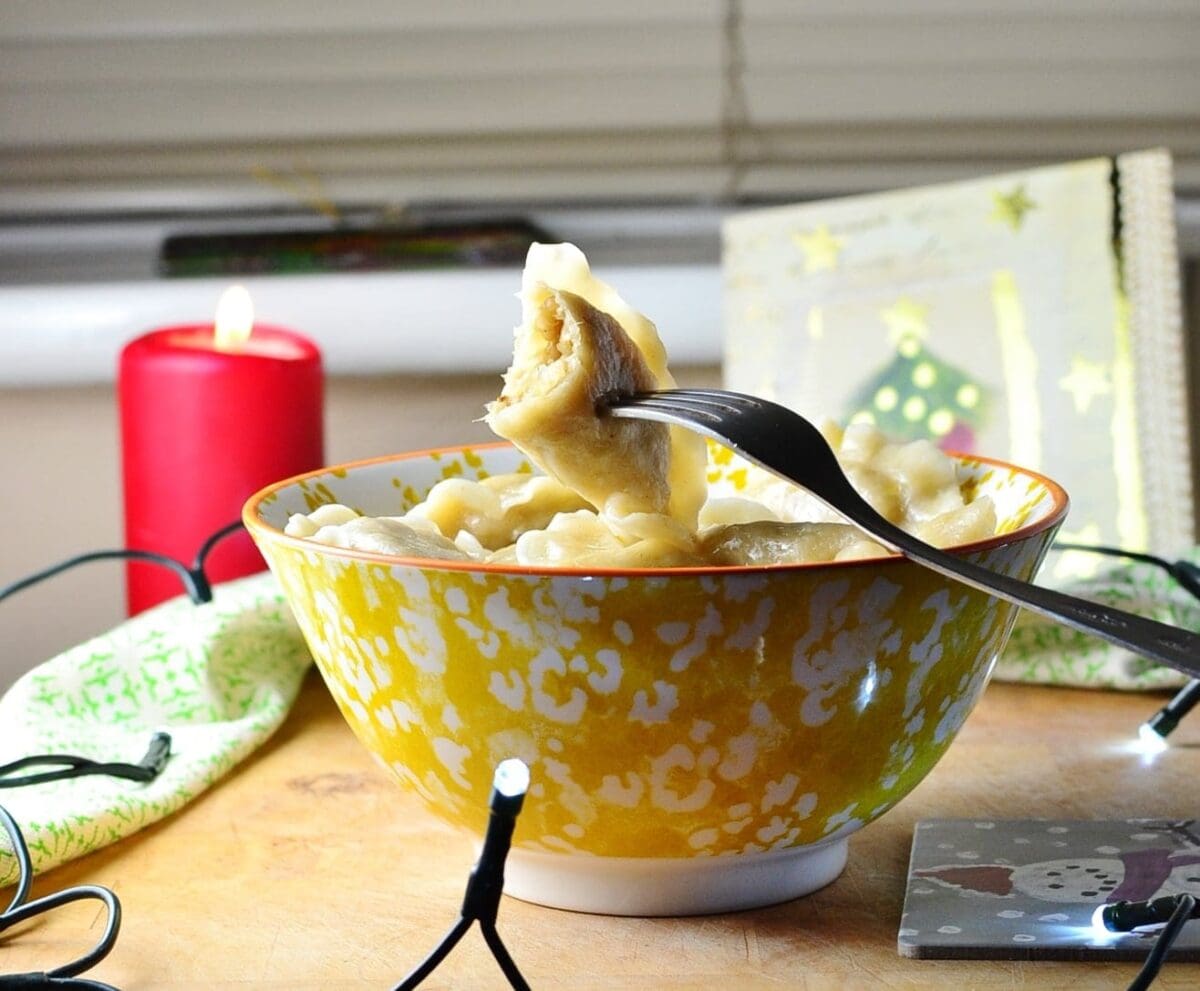 Sauerkraut pierogi in white-and-yellow patterned bowl with one dumpling on fork, Christmas card, lights, red candle and green cloth in background.