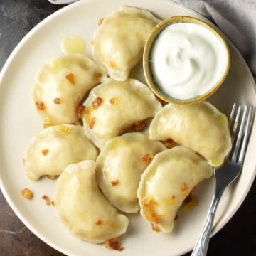 Top down view of cooked potato and cheese pierogi ruskie with sour cream and fork on beige plate.