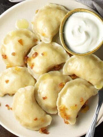 Top down view of cooked potato and cheese pierogi ruskie with sour cream and fork on beige plate.