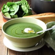 Side view of creamy spinach soup in green bowl with spoon on top of white plate, with spinach, baguette and green cloth in the background.