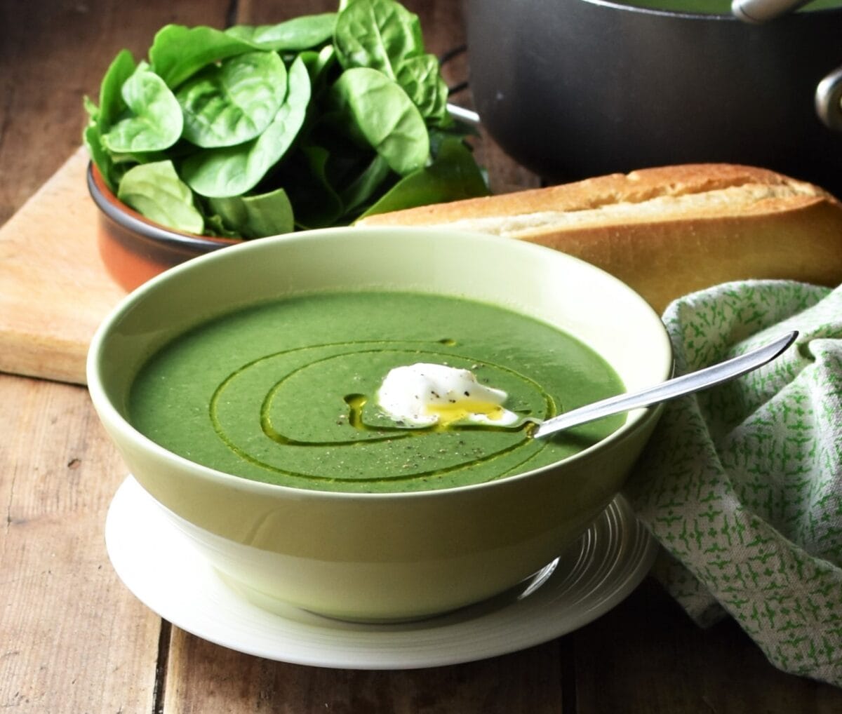 Side view of creamy spinach soup in green bowl with spoon on top of white plate, with spinach, baguette and green cloth in the background.