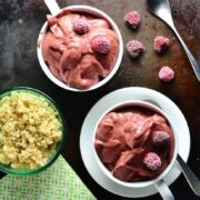 Top down view of raspberry smoothie in 2 white cups, one on saucer with spoon and frozen raspberries, with cooked quinoa in green dish and green cloth in bottom left corner.