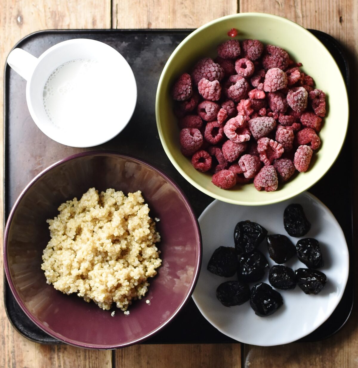 Cooked quinoa in purple bowl, frozen raspberries in yellow bowl, prunes on white saucer and milk in cup.