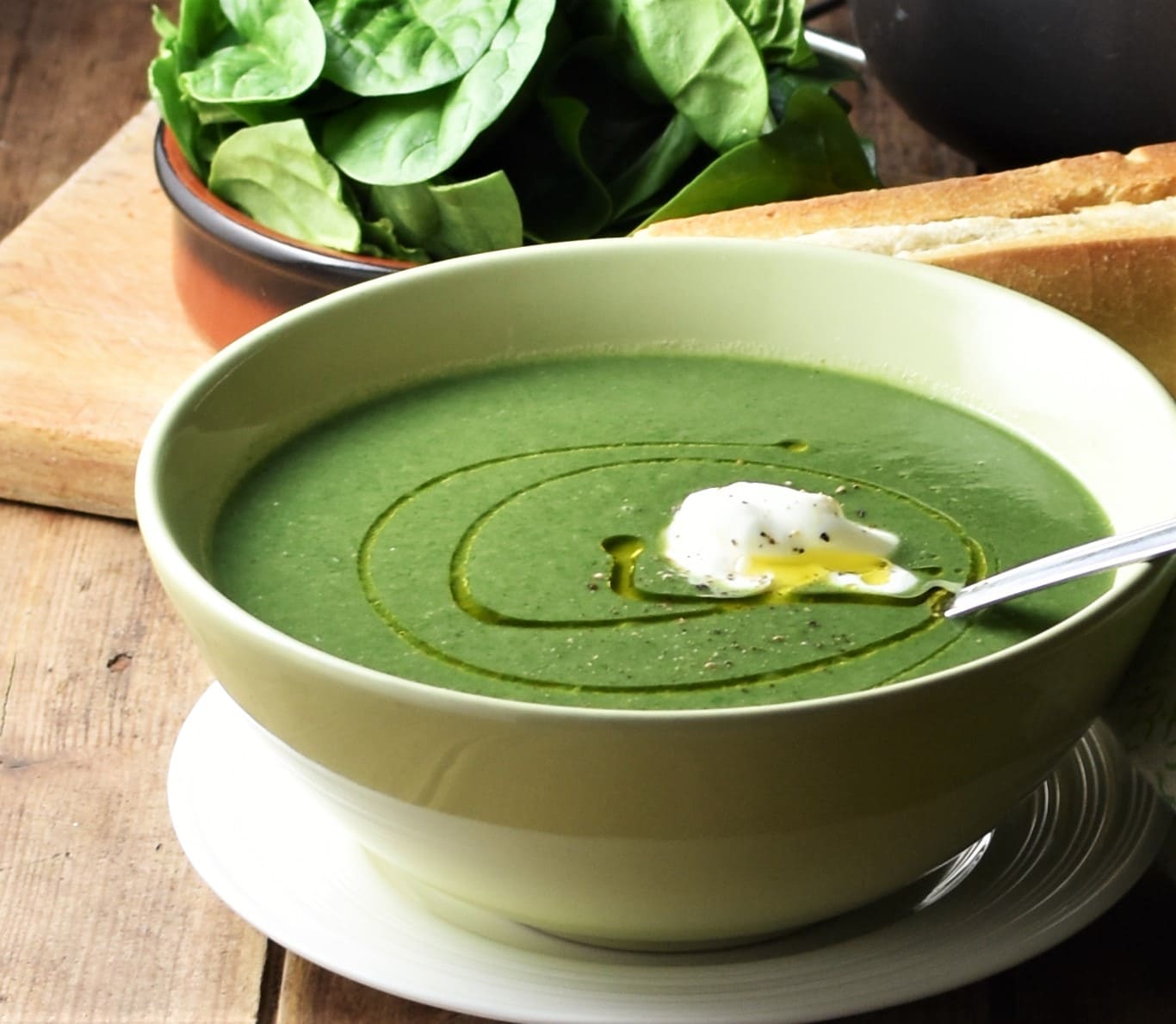 Side view of creamy spinach soup with yogurt in green bowl with spoon on white plate, with spinach in brown dish in background.