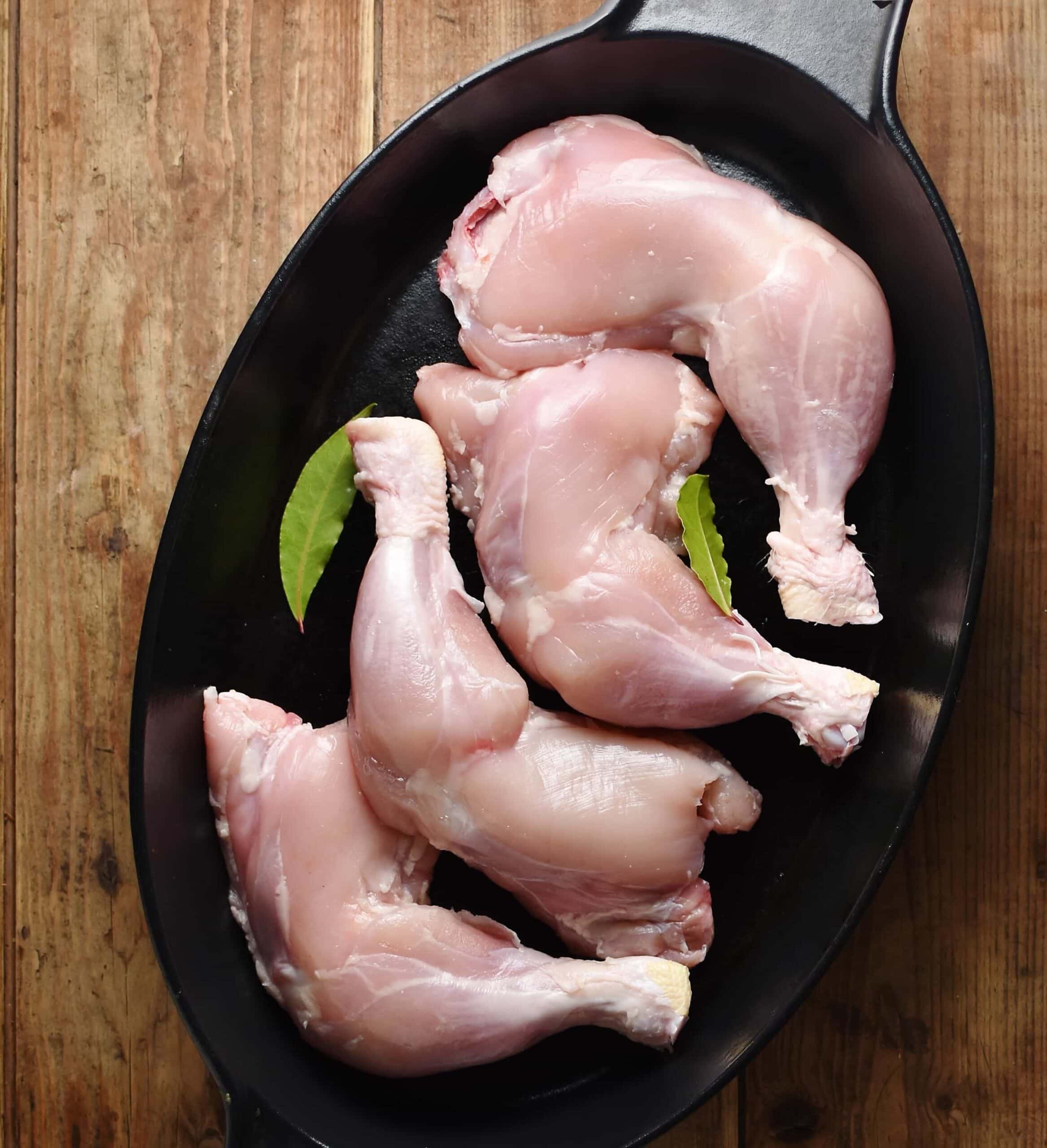 4 skinless chicken legs with bay leaves in black oval dish.