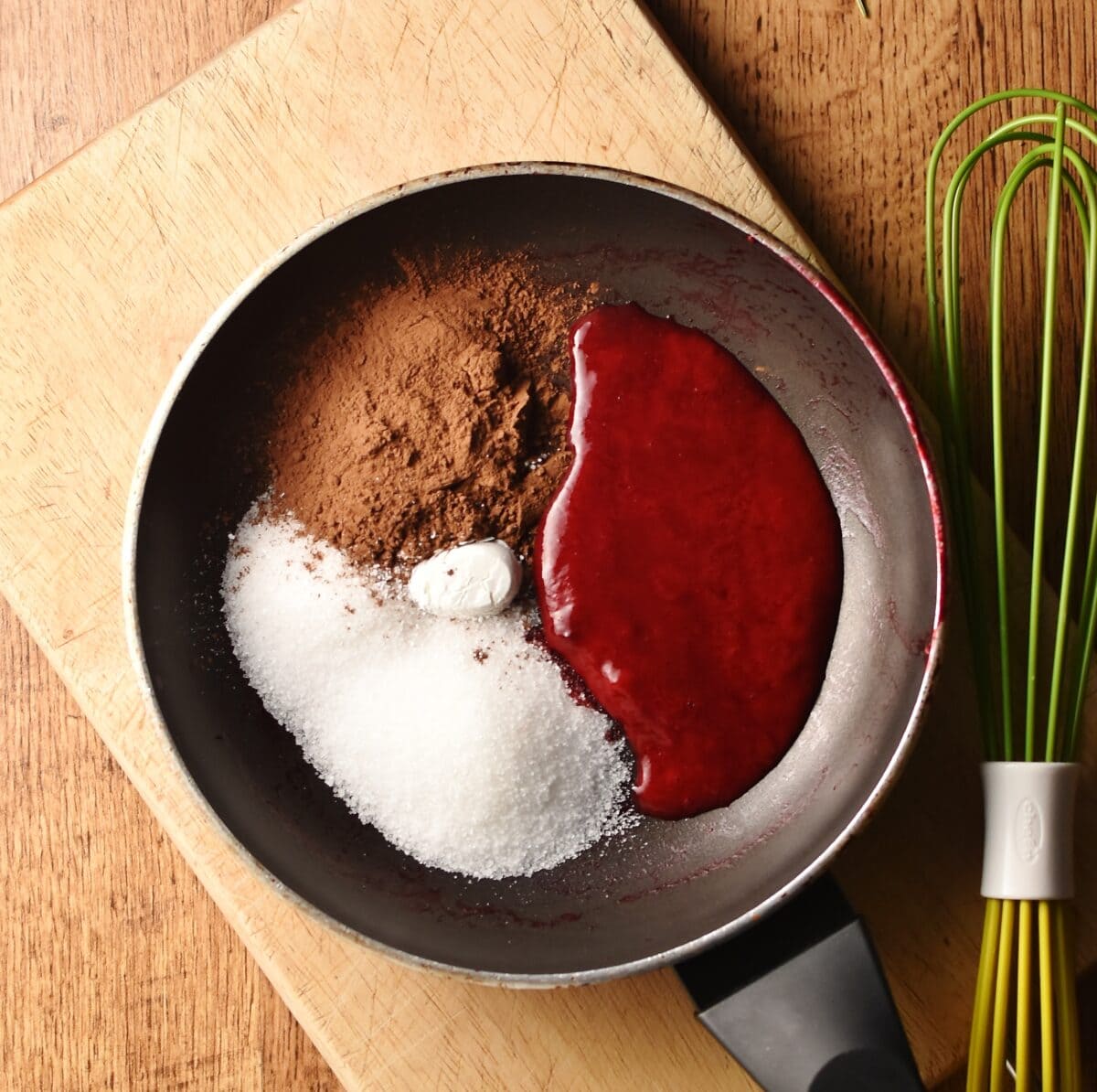 Raspberry sauce, cocoa and sugar in pan on top of wooden board with green whisk to the right.