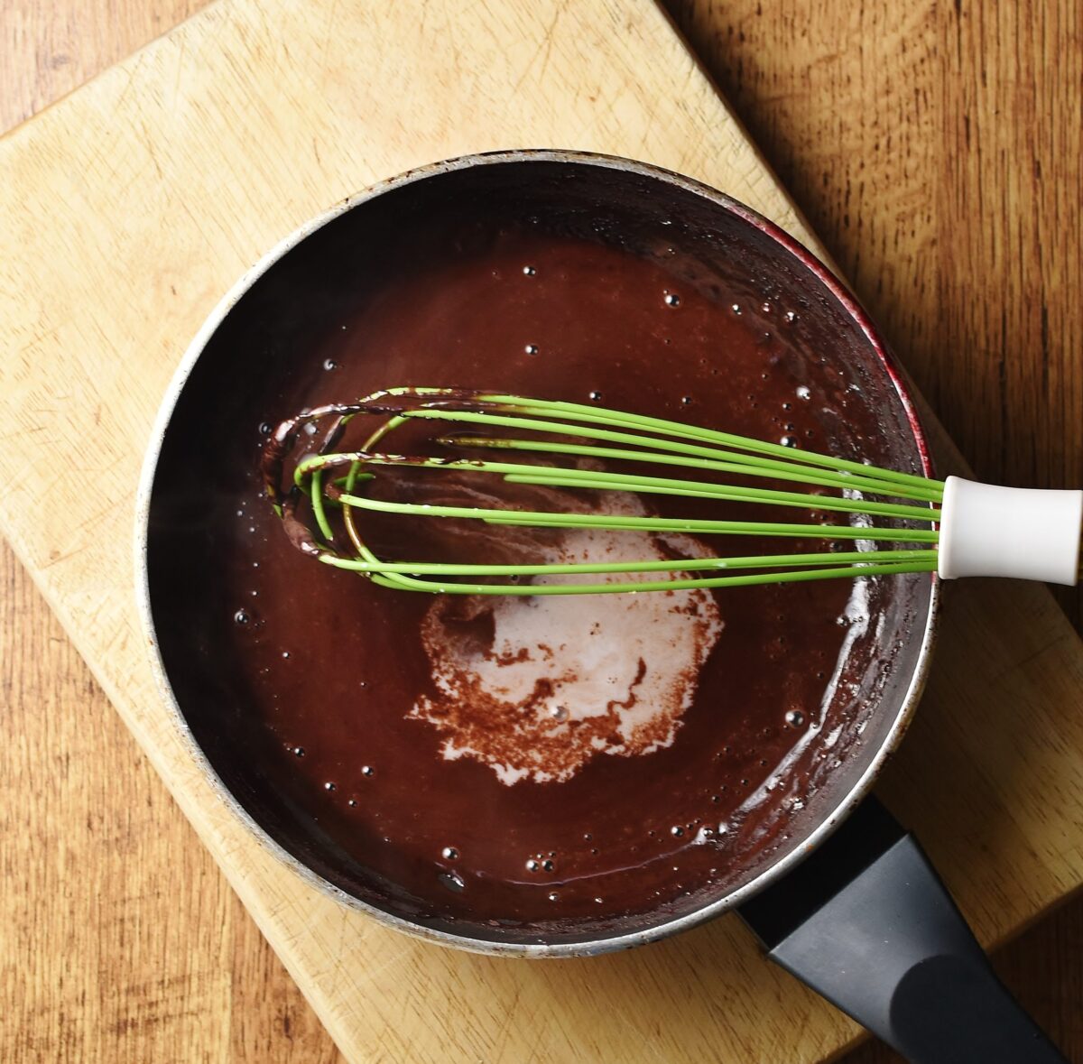 Chocolate sauce with milk visible and green whisk in pan on top of wooden board.