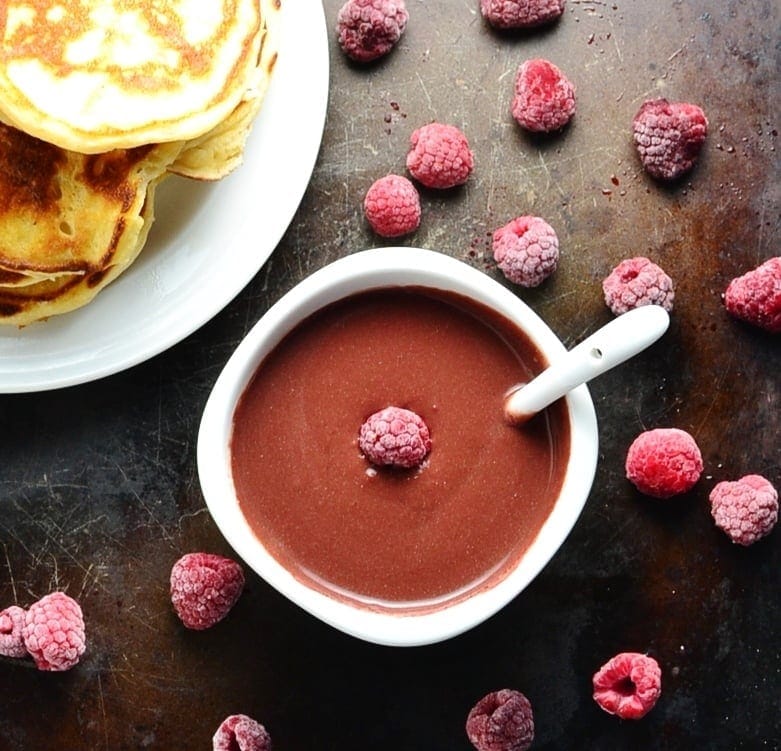 Chocolate sauce with raspberries in white bowl with spoon, with partial view of white plate with pancakes on dark surface.