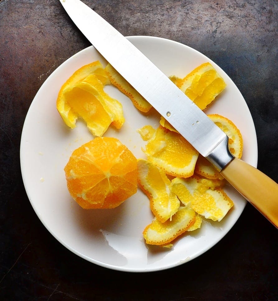 Peeled orange with peel and knife with yellow handle on white plate.
