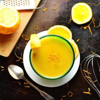 Orange sauce in bowl on white plate with cutting board, oranges, zester, spoon, whisk and orange zest on dark brown surface.