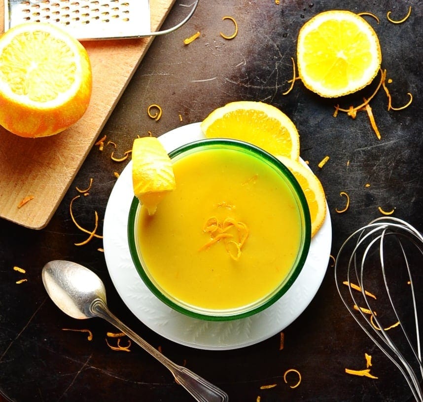 Orange sauce in bowl on white plate with cutting board, oranges, zester, spoon, whisk and orange zest on dark brown surface.