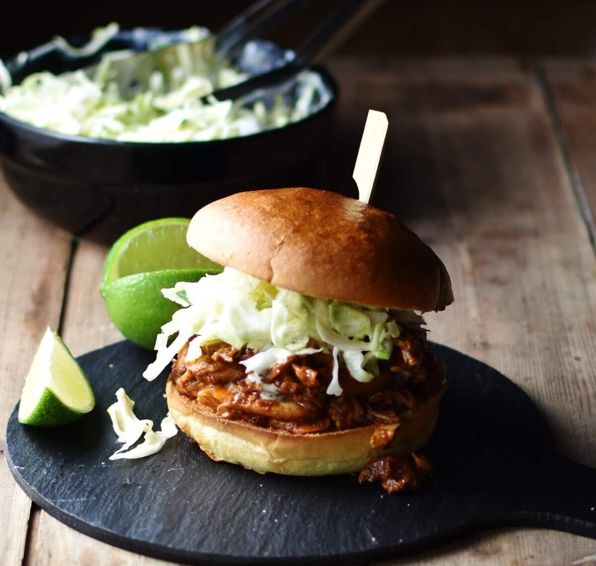 Side view of pulled chicken with coleslaw inside burger bun pierced with bamboos stick, with lime and coleslaw in black dish in background.