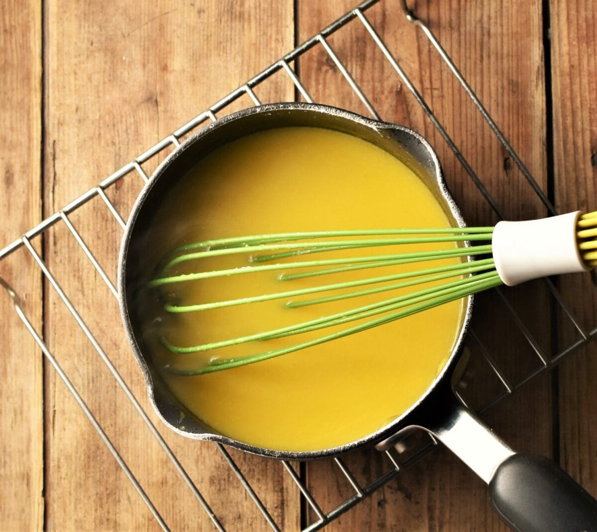 Orange sauce in small pot with green whisk on top of rack.
