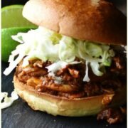 Side view of shredded chicken with coleslaw inside burger bun pierced with bamboos stick, with lime and coleslaw in black dish in background.
