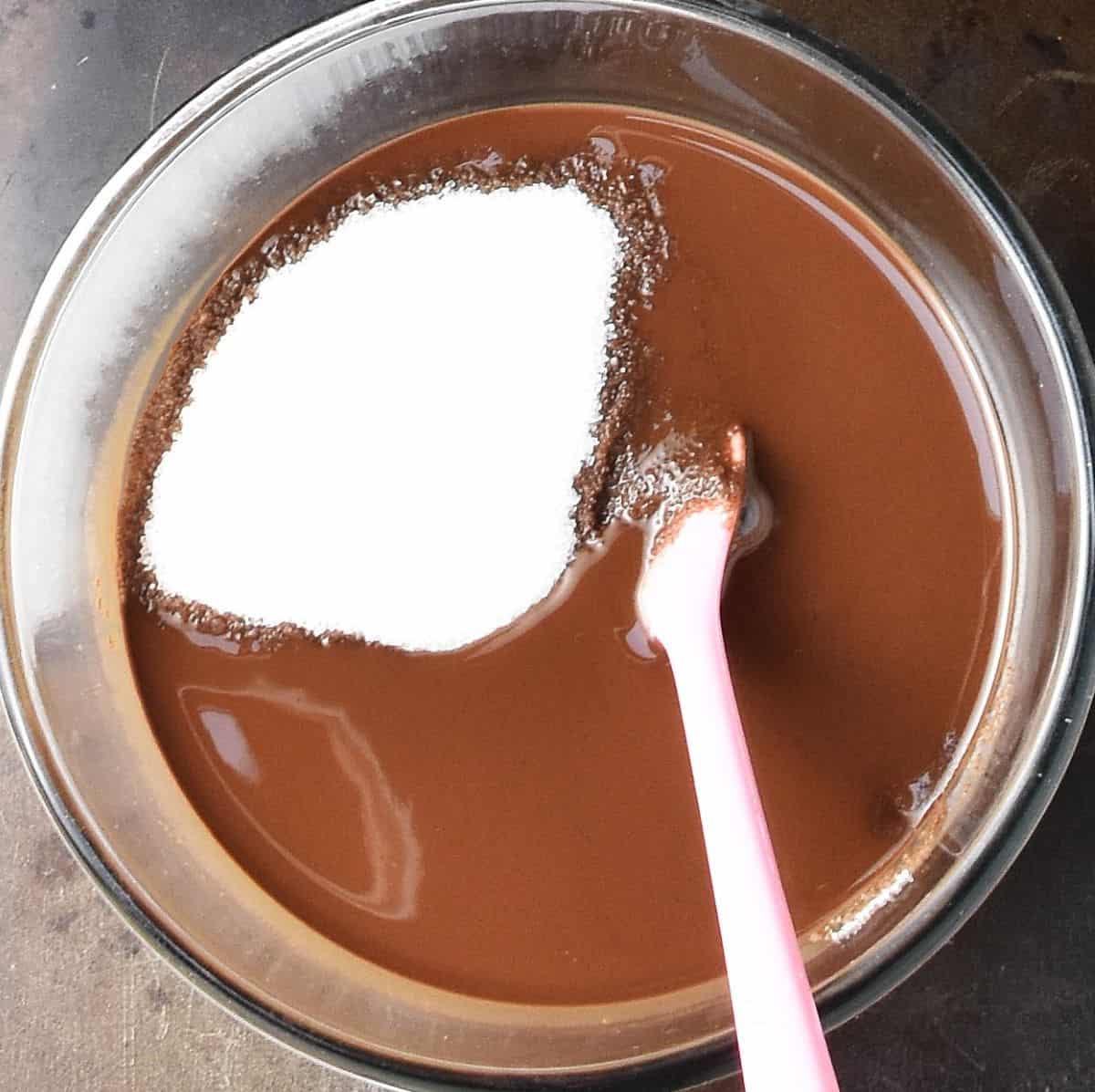 Top down view of melted chocolate with sugar and pink spatula in glass bowl.
