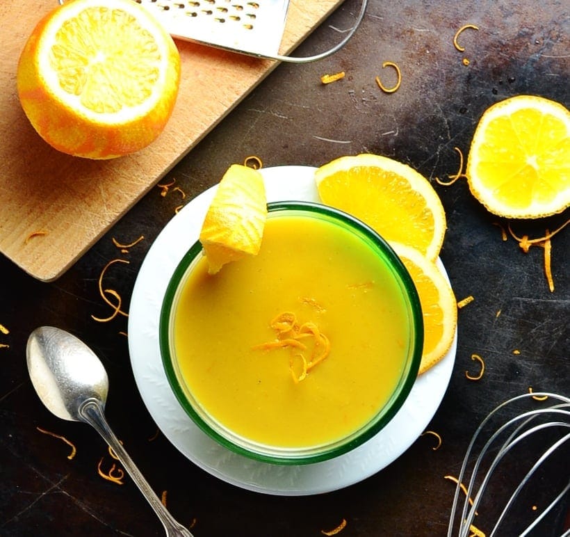 Orange sauce in bowl on white plate with cutting board, oranges, zester, spoon, whisk and orange zest on dark brown surface.