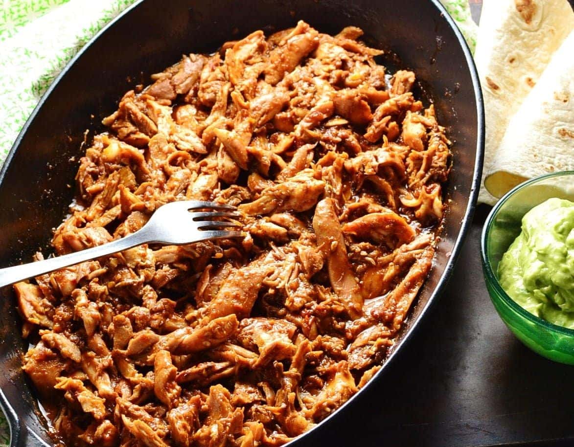 Pulled chicken in black oval cast iron dish with fork, guacamole in small dish, wraps and green cloth.