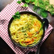 Top down view of chicken noodle soup in black bowl with spoon on red-and-white checkered cloth with dill and parsley on cutting board in top right corner.