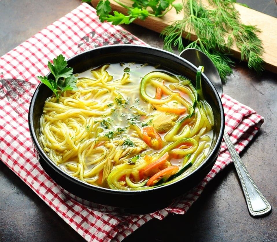 Chicken noodle soup in black bowl with spoon on red-and-white checkered cloth with dill and parsley on cutting board in top right corner.