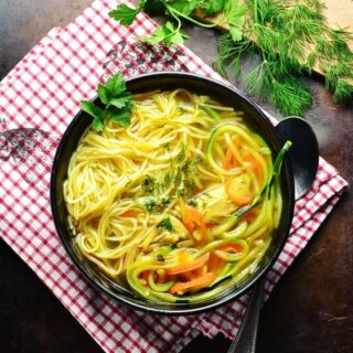 Top down view of chicken noodle soup in black bowl with spoon on red-and-white checkered cloth with dill and parsley on cutting board in top right corner.