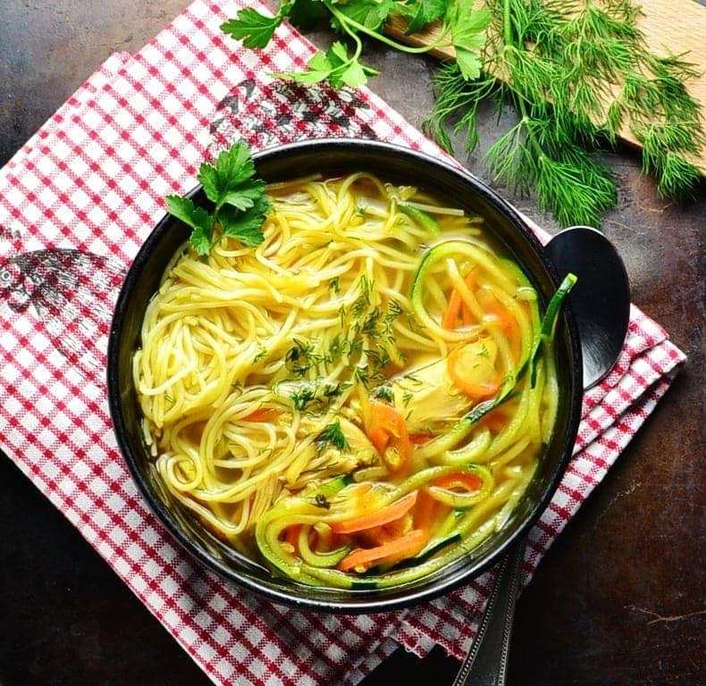 Top down view of chicken noodle soup in black bowl with spoon on red-and-white checkered cloth with dill and parsley on cutting board in top right corner.