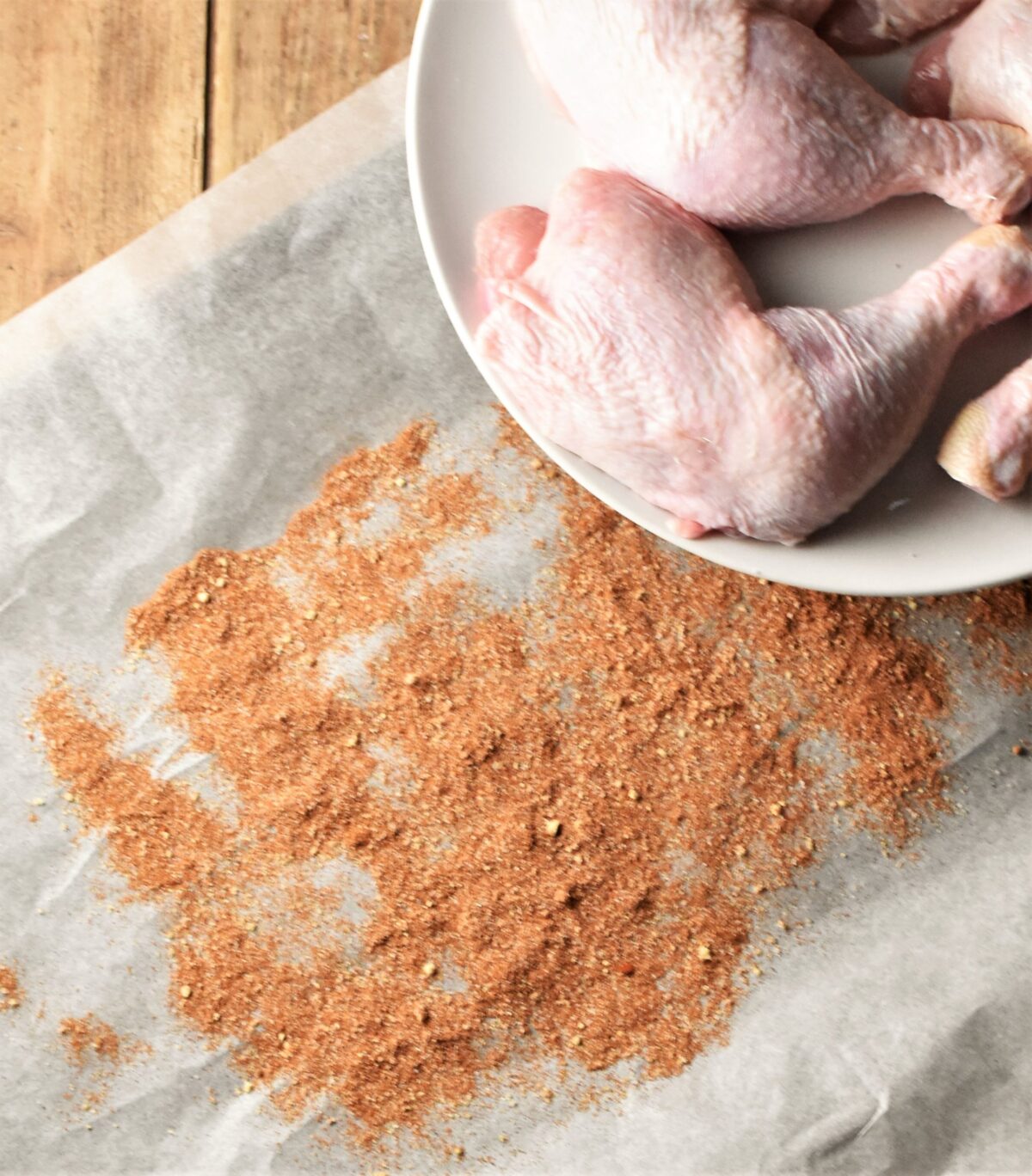 Spice mixture spread over parchment paper and raw chicken leg quarters on white plate in top right corner.