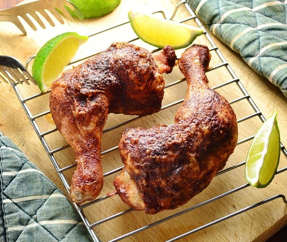 Top down view of 2 crispy chicken leg quarters on top of wire rack with lime wedges and tongs in background.