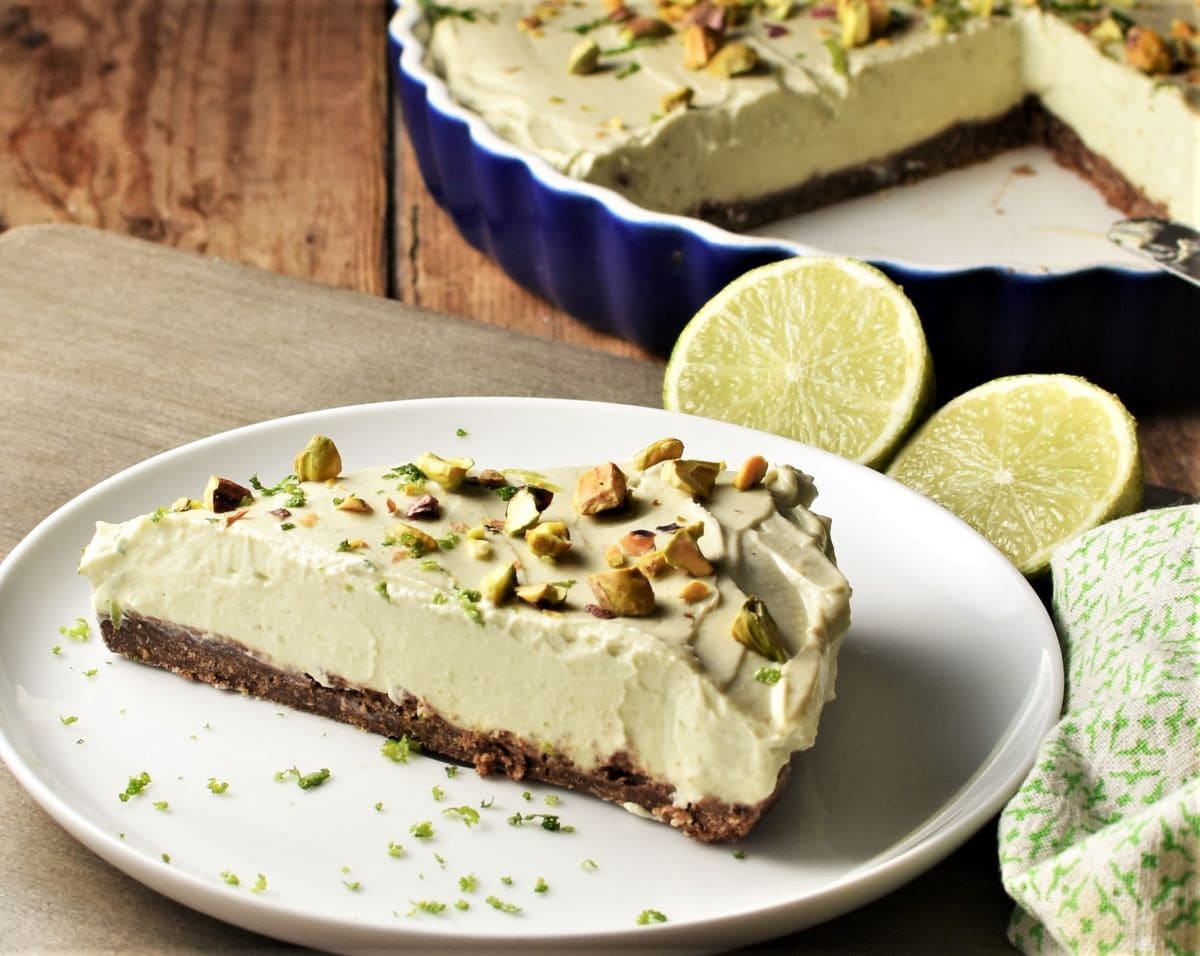 Side view of cheesecake slice with pistachios on plate, with limes in background.