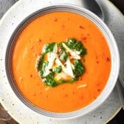 Top down view of creamy res pepper tomato soup with cheese and herbs in grey bowl.