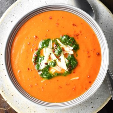 Top down view of creamy res pepper tomato soup with cheese and herbs in grey bowl.