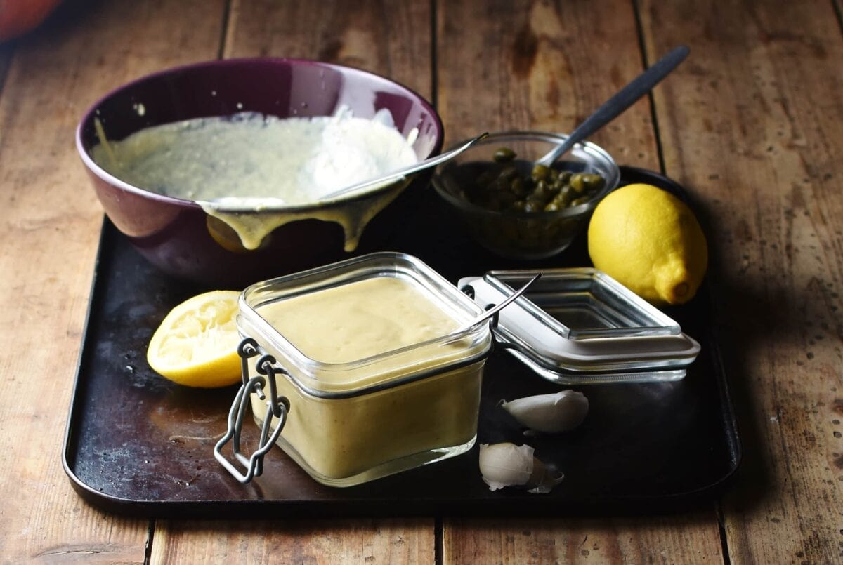 Caesar salad dressing in square open jar with spoon, lemon, dressing in purple bowl and capers in small dish with spoon in background.