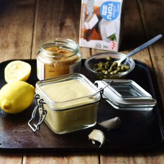 Dressing in square open jar with spoon, lemon, miso paste in jar, capers in small dish with spoon and silken tofu package in background.