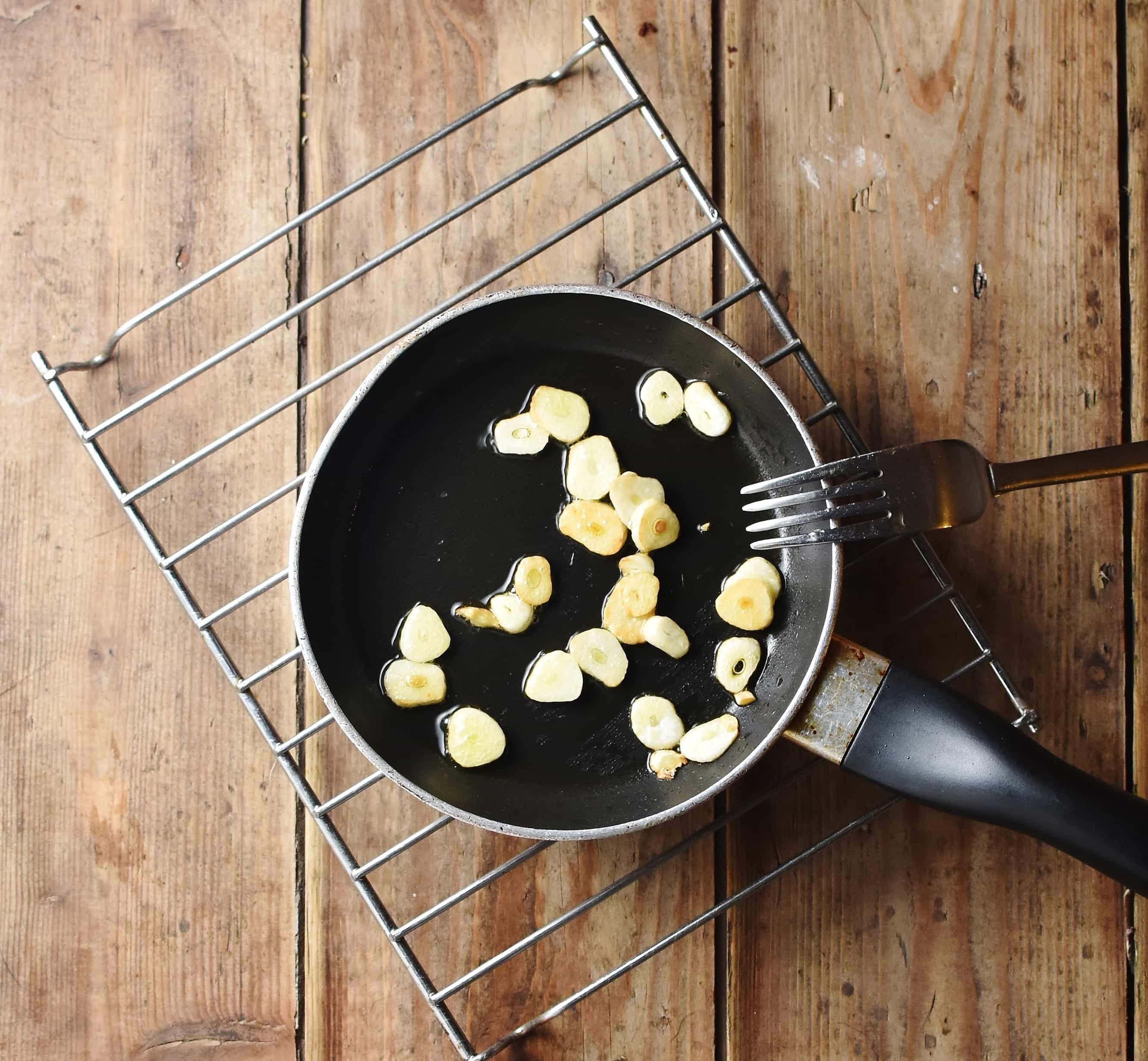 Slices of garlic in small pan with fork on rack.