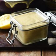 Caesar dressing in square open jar with spoon, lemon and dressing in purple bowl in background.
