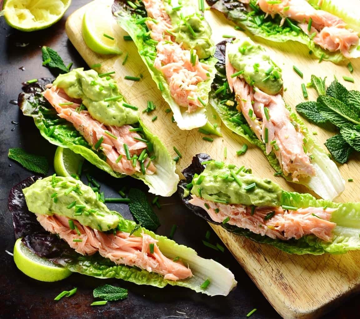 Lettuce wraps with salmon and guacamole with garnish of herbs and lime wedges on top of wooden board and dark table.