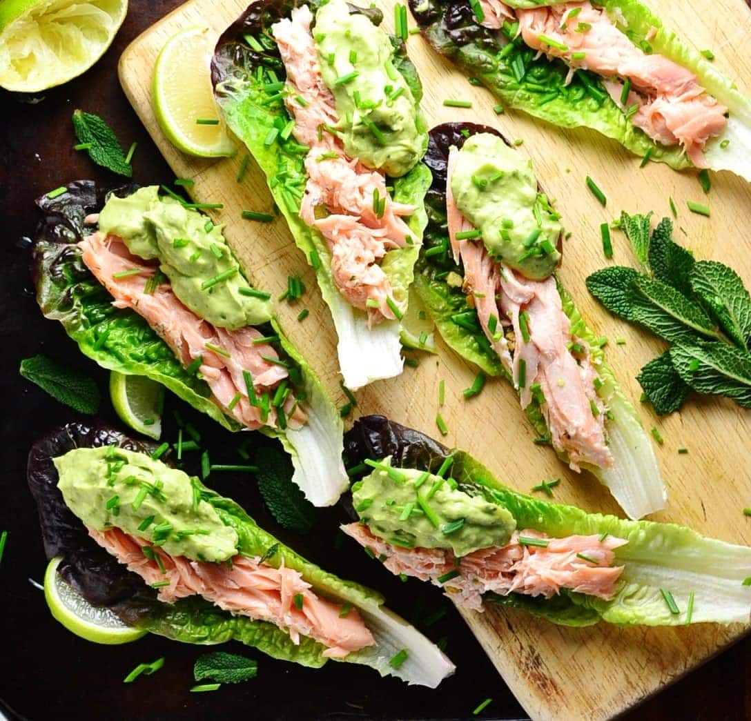 Top down view of lettuce wraps with salmon and avocado sauce with lime wedges and garnish of herbs on top of wooden board and dark table.