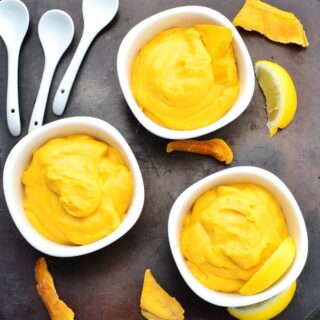 Top down view of yellow coloured vegan mango mousse in 3 white bowls with dried mango and 3 white spoons on dark surface.