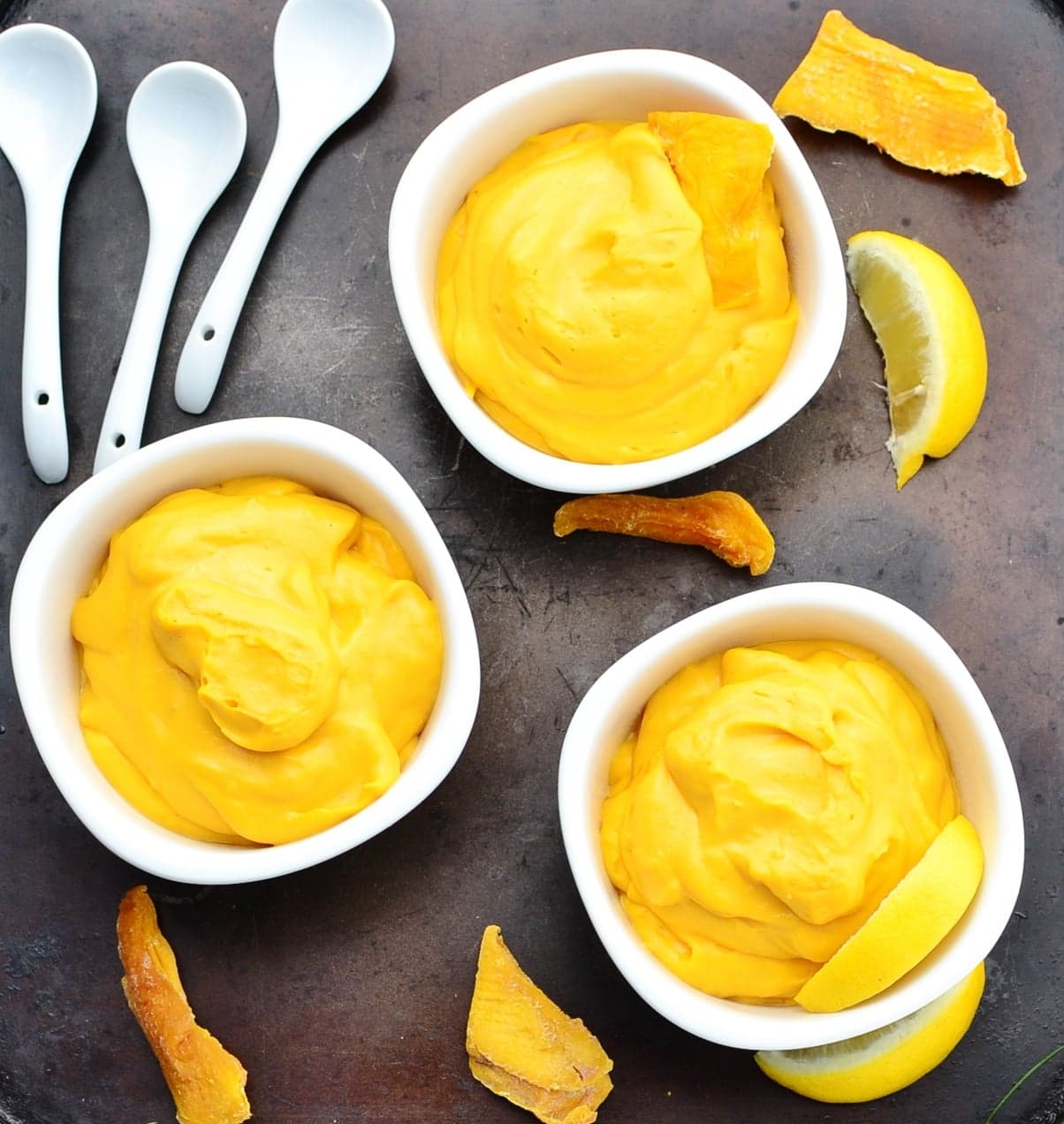 Top down view of yellow coloured mousse in white bowls with dried mango pieces, lemon wedges and 3 white spoons on dark surface.