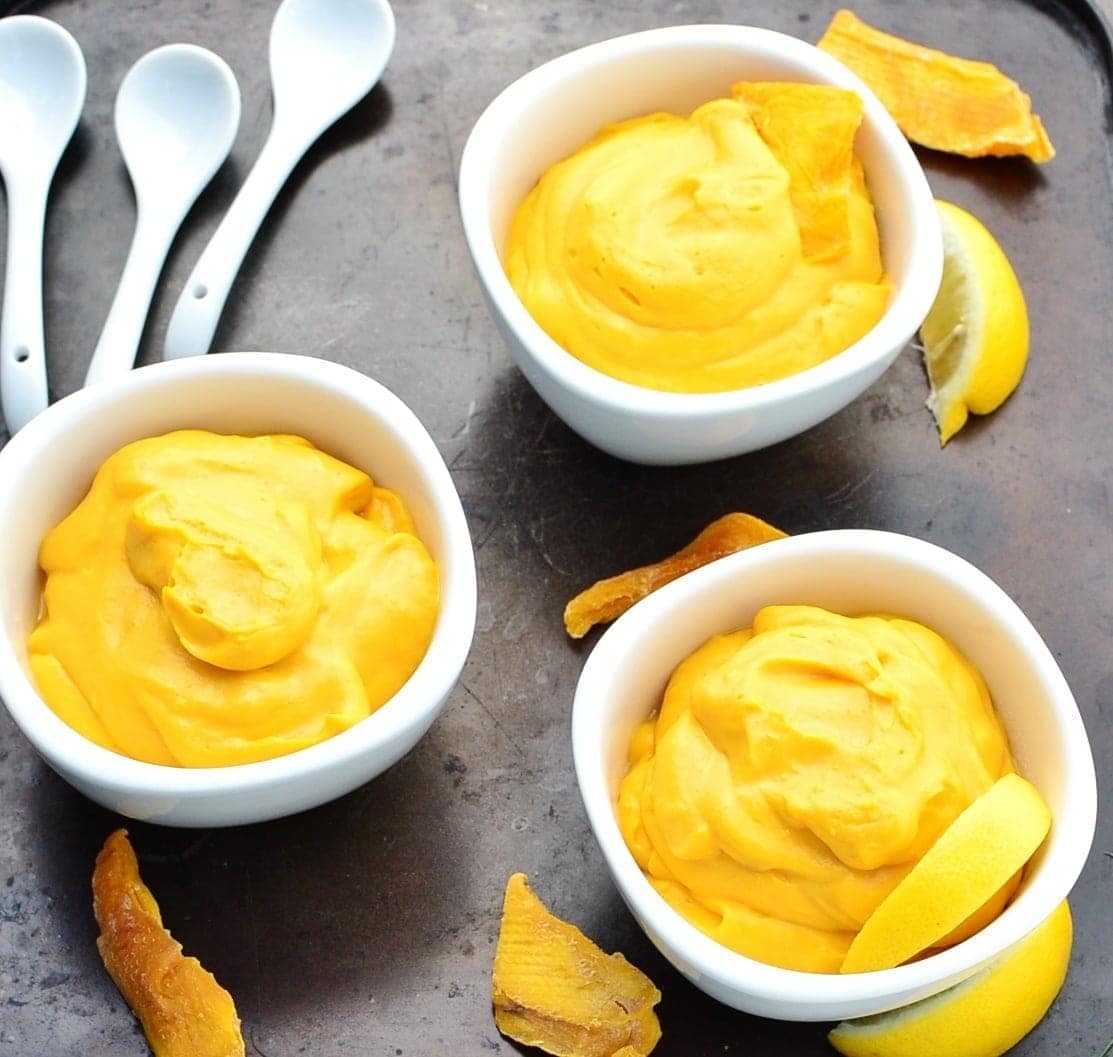 Top down view of yellow mango mousse in 3 white bowls with dried mango and 3 white spoons on dark surface.