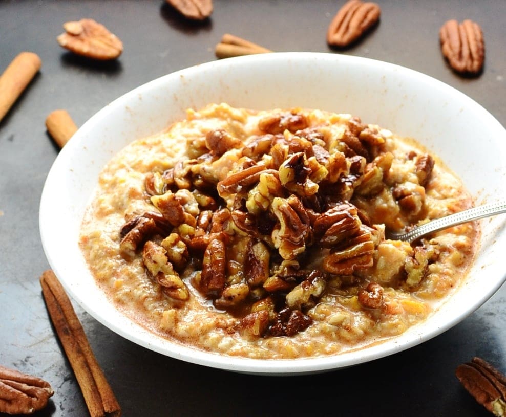 Side view of sweet potato overnight oats with pecans and spoon in white bowl on top of dark table with cinnamon sticks and pecans scattered about.