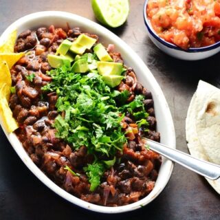 Black bean stew in white oval dish garnished with chopped cilantro, avocado and nachos, with lime, salsa in blue dish and tortilla on dark brown surface.