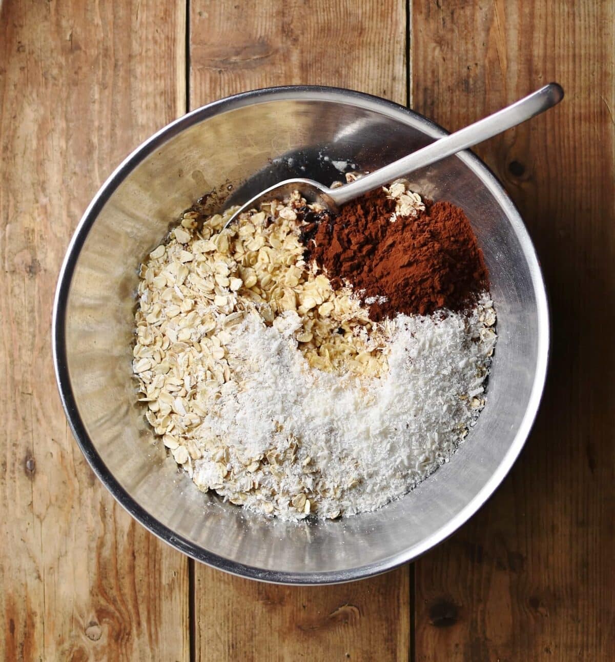 Oats, cocoa and coconut with spoon inside metal bowl.