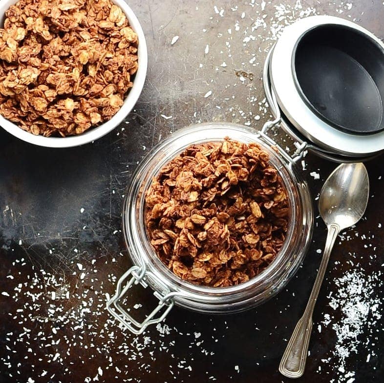 Top down view of chocolate granola inside open jar, with spoon, coconut flakes and granola in white bowl.