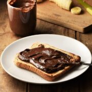 Side view of toast with chocolate hazelnut spread and knife on white plate, with spread in cup and cutting board in background.